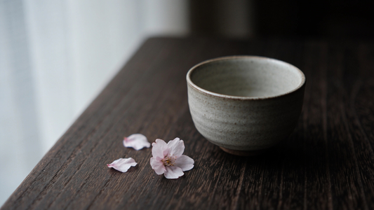 Teetasse mit Kirschblütenblättern auf dunklem Holz – Mono no Aware, die Schönheit des vergänglichen Moments; KI-gestützt erstellt